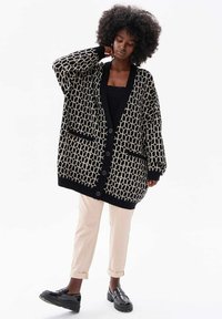 Oversized zwart en crèmekleurig vest met geometrisch patroon, knoopsluiting en twee voorzakken, gecombineerd met lichte broek en schoenen.