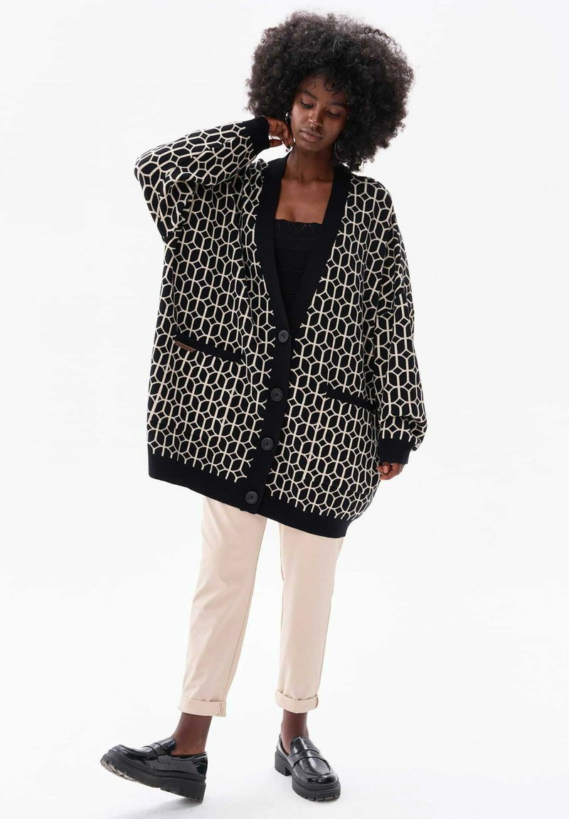 Oversized zwart en crèmekleurig vest met geometrisch patroon, knoopsluiting en twee voorzakken, gecombineerd met lichte broek en schoenen.