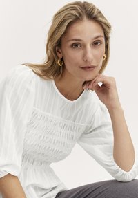 Vit, texturerad blus med puffärmar och rynkad detalj längs bodicen, buren med grå byxor. Guldörhängen som accent.