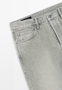 Lichtgrijze spijkerbroek met voorzak, riemlussen en metalen knoopsluiting, plat gelegd op een witte achtergrond.