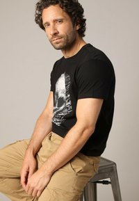 T-shirt noir en coton avec un graphisme de crâne et du texte blanc. Assorti à un pantalon cargo beige, affichant une coupe décontractée et un style casual.
