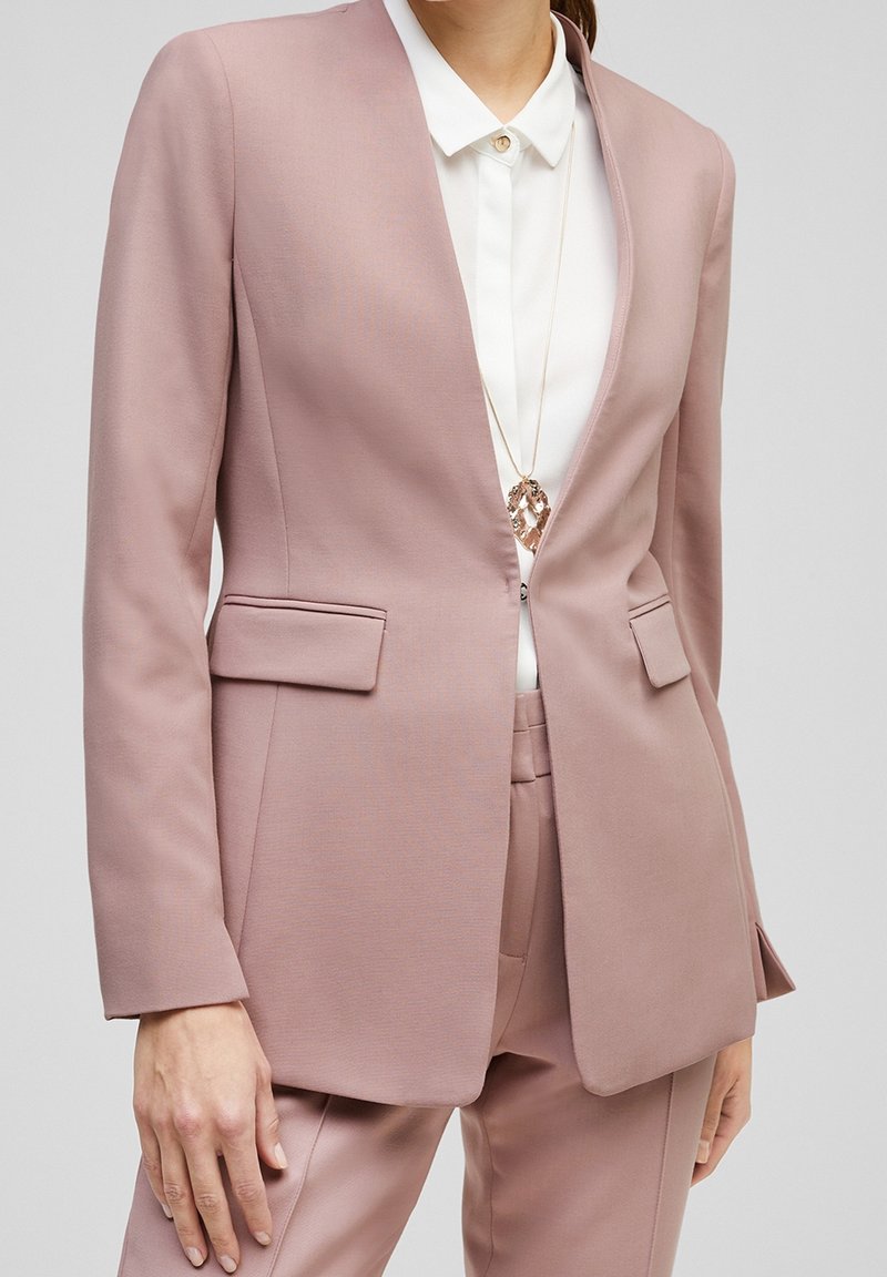 Ljust rosa skräddarsydd kavaj med två framfickor, slagkrage och slät textur. Bärs över en vit skjorta med en guldk knapp.