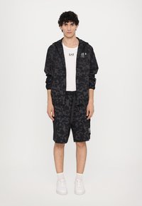 Svart treningsantrekk med en zip-up jakke i leopardmønster og matchende shorts. Begge delene har detaljer i en lysere nyanse.