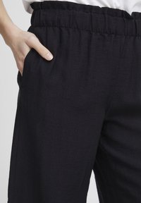 Zwarte, lichte broek met een elastische tailleband en zijzakken. De stof heeft een gestructureerde afwerking, wat subtiele details toevoegt.