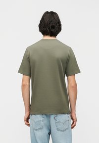 Άνδρας με σκούρα σγουρά μαλλιά, που φοράει ένα απλό ελιά πράσινο κοντομάνικο μπλουζάκι και ανοιχτό μπλε τζιν, στέκεται με την πλάτη προς τα εμπρός σε λευκό φόντο.