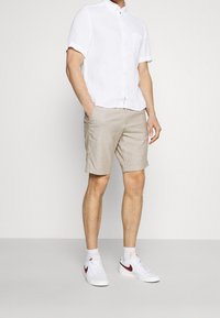 Camicia di lino bianca a maniche corte, pantaloni beige e sneakers bianche con dettagli bordeaux. La camicia ha una chiusura con bottoni e una tasca sul petto.