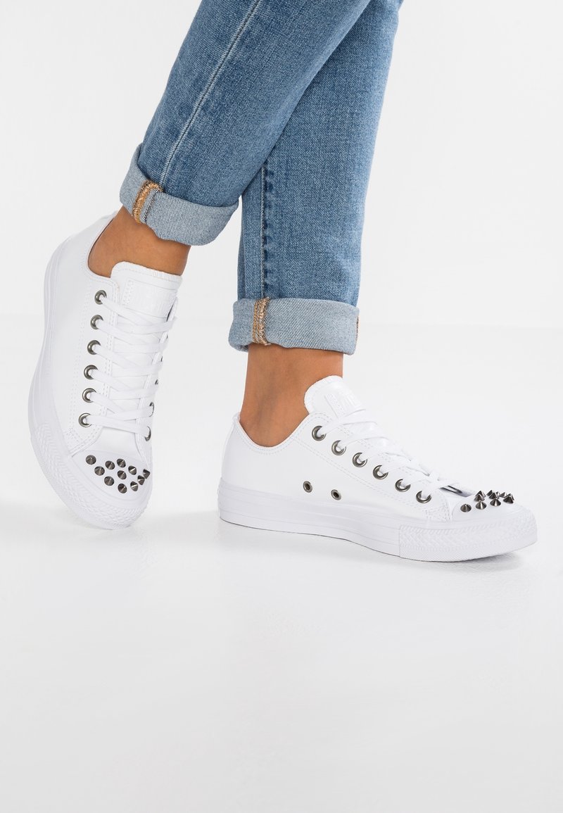 White canvas sneakers with rounded toe, metallic studs on the front, and silver eyelets for laces. Paired with rolled-up blue denim jeans.