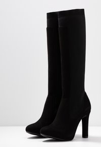 Bottes noires à talons hauts avec une finition en daim, design montantes jusqu'au genou et haut en tricot. Le talon courbé ajoute une silhouette structurée.