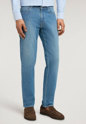 Hombre vistiendo vaqueros de mezclilla azul claro, camisa azul clara y mocasines de ante marrones, de pie contra un fondo claro liso.