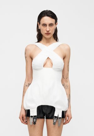 Γυναίκα που φοράει λευκό τοπ με χιαστί λεπτομέρεια και άνοιγμα, μαύρο σορτς και έχει πολλά τατουάζ στα χέρια της, μπροστά σε απλό φόντο.