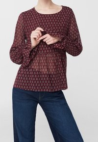 Blusa trasparente color bordeaux con lunghe maniche a volant, caratterizzata da un motivo ripetitivo in piccolo bianco. Abbinata a jeans a gamba larga blu scuro.