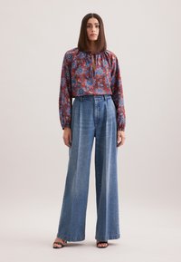 Blusa floreale rossa con maniche lunghe e colletto allacciato, abbinata a jeans in denim blu a vita alta e gamba larga. Tessuto morbido e vestibilità ampia.