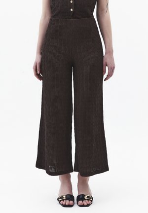 Kobieta ubrana w teksturowane, ciemnobrązowe szerokie spodnie i czarne sandały na otwartym palcu ze złotymi klamrami, stojąca na białym tle.