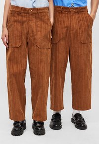 Calças de cotim castanhas com riscas verticais, apresentando bolsos laterais, fecho com botão e um corte descontraído. Usadas com calçado preto.