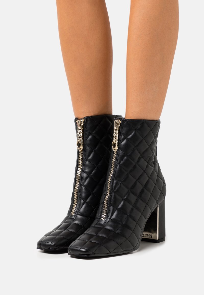 Bottes en cuir noir matelassé à la cheville avec des fermetures éclair avant dorées et des talons blocs portées sur des jambes nues.