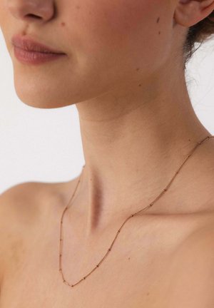 Close-up of a woman's neck and collarbone wearing a delicate gold chain necklace against a neutral background.