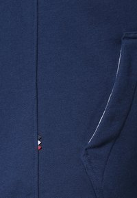 Mörkblå sweatshirttyg med en slät textur, som har en sidoficka och en liten flerfärgad logodetalj nära fållen.