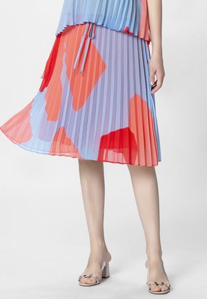 Jupe midi plissée lavande ornée de grandes formes géométriques rouges et orange, portée avec des sandales à talons blocs à brides transparentes.