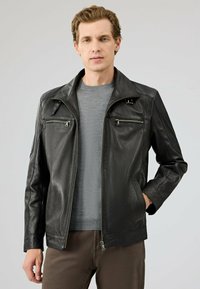 Zwarte leren jasje met een gestructureerde afwerking, ritszakken en een kraag. Gedragen over een grijze trui en een paarse broek.