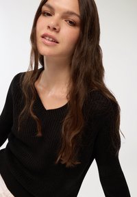 Czarny sweter z prążkowanego materiału z dekoltem w kształcie litery V, z długimi rękawami i teksturowanym wykończeniem. Modelka ma luźne fale w włosach.