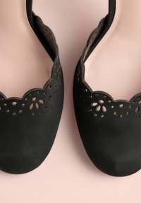 Sorte ruskindssko med bølget kant og laserskårne blomsterbelægninger på tåområdet, der har en glat tekstur og rund form.