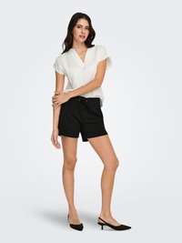 Witte kortärmelig top met een V-hals, gecombineerd met zwarte korte broek. Model draagt zwarte slingback schoenen met spits toelopende neus, tegen een effen achtergrond.