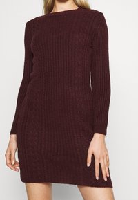 Femme portant une robe pull en tricot côtelé bordeaux à manches longues, debout avec les bras détendus devant un fond clair uni.