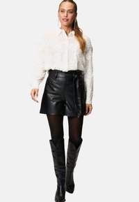 Chemisier blanc avec broderies florales, shorts en cuir noir à enrouler, collants noirs transparents et bottes noires montantes à bouts pointus.