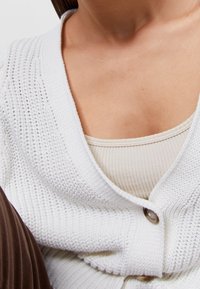 Bílý pletený svetr s hlubokým výstřihem do V a detaily knoflíků. Vnitřní vrstva je světle béžový tank top s texturou.