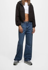 Czarna krótka kurtka z kapturem i elastycznymi mankietami, zestawiona z jasnoniebieskimi dżinsami o szerokich nogawkach oraz szarymi sportowymi butami. Prosty, codzienny strój na zewnątrz.