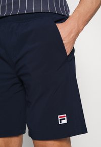 Marineblå shorts laget av lett materiale, med elastisk midjebånd, sidelomme og et teksturert logo-patch med røde og hvite detaljer.