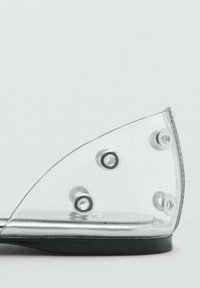 Transparent flat shoe with a pointed toe, chrome-accented rim, clear upper, and black sole. Features circular metal details.