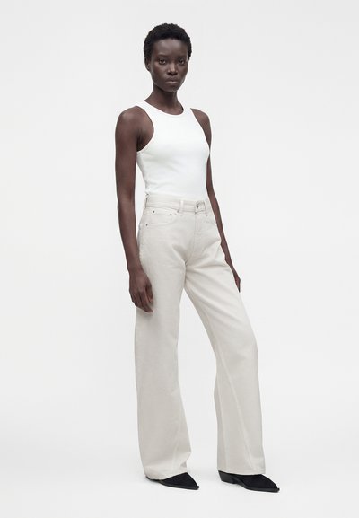 Femme debout portant un haut blanc sans manches, un pantalon large beige clair et des chaussures noires pointues sur un fond clair uni.