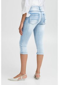 Pantalones capri de mezclilla azul claro con dobladillos enrollados, dos bolsillos traseros y una textura suave, combinados con zapatos blancos puntiagudos.