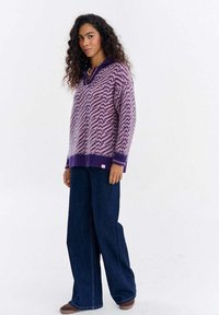 Kobieta o długich kręconych włosach, ubrana w fioletowo-różowy wzorzysty sweter z kołnierzem, szerokie ciemnoniebieskie spodnie i brązowe buty, stojąca bokiem.