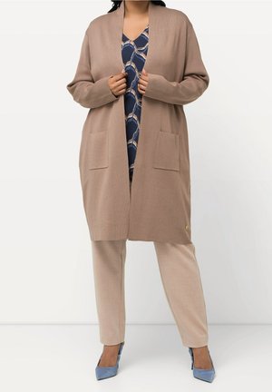 Kvinde iført beige lang cardigan med frontlommer, beige bukser, blå mønstret top og blå spidse hæle, stående på hvid baggrund.