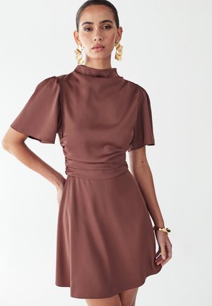 Kvinde iført en brun satin kortærmet kjole med høj krave, accessoriseret med store gyldne blomsterøreringe og et gyldent armbånd.