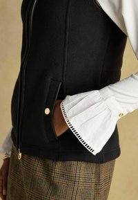 Čierny fleecový vest so zipsom vpredu, umelou koženou obrubou, bočnými vreckami so zlatými akcentmi a bielou košeľou s detailom rozšíreného manžetu.