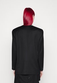 Černý blazer s přiléhavým střihem, s upravenými ramenními partiemi, dlouhými rukávy a knoflíkovými detaily na manžetách. Hladká textilie.