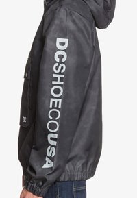 Black jacket with a hood, featuring a prominent gray logo "DCSHOECOUSA" down the left sleeve and elastic cuffs. Smooth, water-resistant fabric.