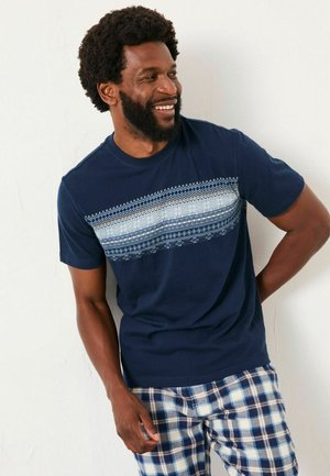 Lächelnder Mann mit Bart, der ein marineblaues gemustertes T-Shirt und karierte Shorts in Blau und Weiß trägt, vor einer weißen Wand steht.