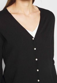 Svart cardigan med V-ringning, med pärlknappar nedtill, gjord av mjukt stickat material med en slät textur.