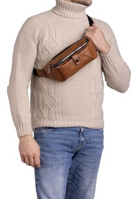 Marsupio in pelle marrone con una texture liscia, cerniera argentata e chiusura a bottone. Indossato diagonalmente da una persona in un maglione di colore crema.