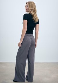 Blonďatá žena stojí, má na sobě černý top s krátkým rukávem a volné šedé kalhoty, dívá se přes rameno na jednobarevném světlém pozadí.