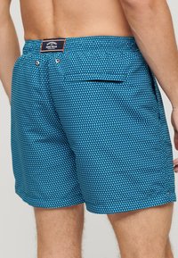Blauwe zwemshorts met een geometrisch driehoekpatroon, een elastische tailleband en een achterzak met een knoop. Merktag zichtbaar op de taille.