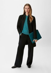 Schwarzer langer Mantel über einem strukturierten, türkisfarbenen Pullover, kombiniert mit schwarzen weit geschnittenen Hosen. Hält eine türkisfarbene Laptop-Hülle. Einfaches, klares Design.