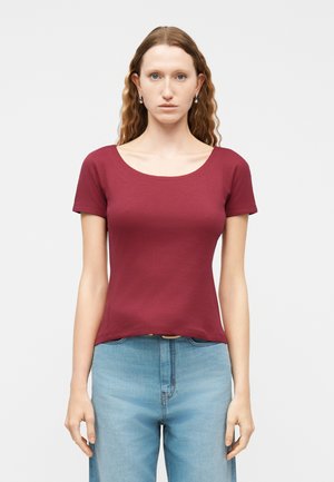 Femme debout, face à l'avant, portant un haut ajusté bordeaux à manches courtes et un jean taille haute bleu clair, devant un fond uni.