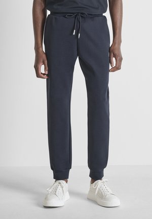 Uomo che indossa pantaloni jogger neri con vita con coulisse e sneakers bianche, in piedi su un pavimento grigio chiaro davanti a uno sfondo bianco.