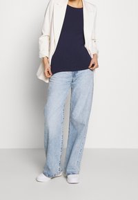 Granatowy top bez rękawów w zestawieniu z białą marynarką, jasnoniebieskimi spodniami o szerokich nogawkach oraz białymi trampkami. Skupienie na czystych liniach i swobodnym kroju.