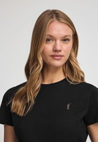 Svart bomull t-shirt med korta ärmar, med en liten guldlogotyp på den övre vänstra sidan. Slät textur, klassisk passform.